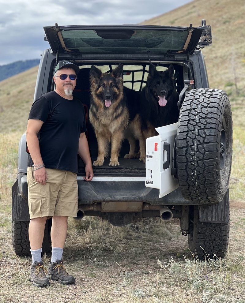 Doug Case standing beside his Jeep on an Idaho hillside with two German Shepherds in the back